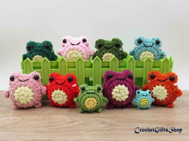 Mehrere bunte, gehäkelte Frosch-Amigurumi aus Plüschgarn in zwei Reihen vor einem grünen Zaun