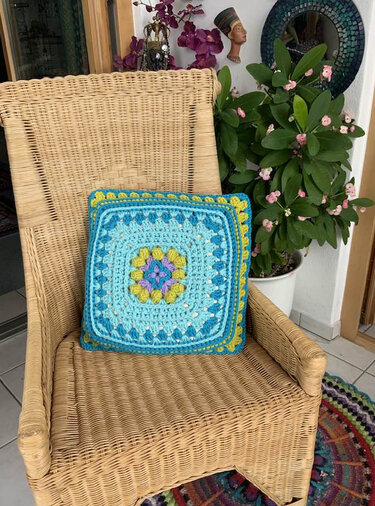 Quadratisches Häkelkissen in Türkis und Blau mit farbigem Granny Square auf einem Korbsessel.