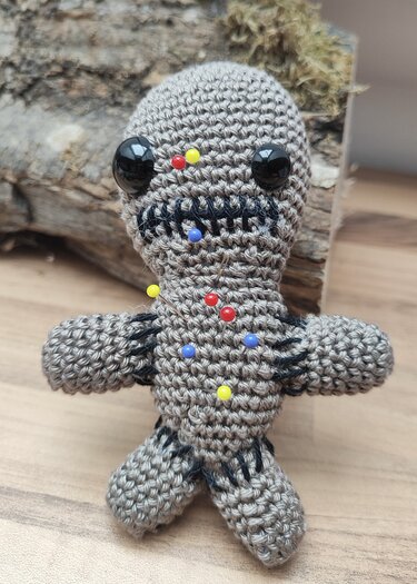 Gehäkelte graubraune Amigurumi-Puppe mit schwarzen Sicherheitsaugen und bunten Stecknadeln