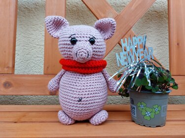 Rosa gehäkeltes Amigurumi-Schwein mit rotem Schal auf einer Holzbank neben einem Topf mit Glücksklee und Happy-New-Year-Schriftzug