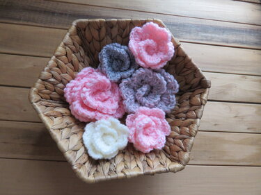 Korb mit gehäkelten Blumen in Rosa, Grau und Creme auf einer Holzoberfläche.