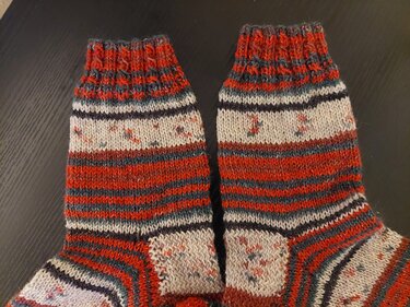 Zwei handgestrickte Socken mit roten, grauen und cremefarbenen Streifen und geripptem Bündchen, liegend auf dunklem Untergrund