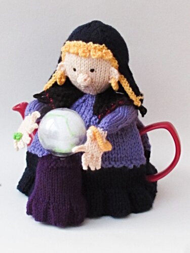Hand-knitted witch tea cozy in black and purple holding a clear decorative sphere on a red teapot.