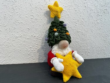 Gehäkelter Weihnachtswichtel mit grünem Tannenhut und gelbem Sternkissen vor weißer Wand
