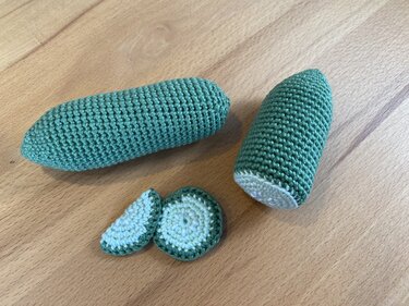 Gehäkelte Gurke mit halbiertem Stück und zwei Scheiben auf einer Holzoberfläche