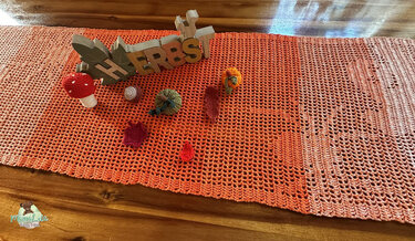 Gehäkelter Tischläufer im Orange-Verlauf mit gehäkeltem Pilz, Kürbissen, Blättern und Holzschrift HERBST auf einem Holztisch.