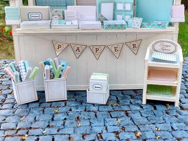 Miniatur-Bastelstand mit pastellfarbenen Designpapieren, Papierrollen und Notizblöcken