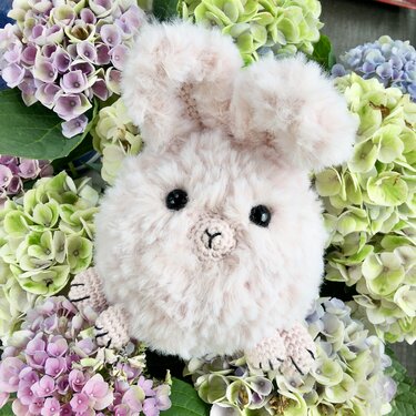 Flauschiger hellrosa gehäkelter Hase mit schwarzen Augen und gestickter Nase zwischen lila-grünen Hortensien.