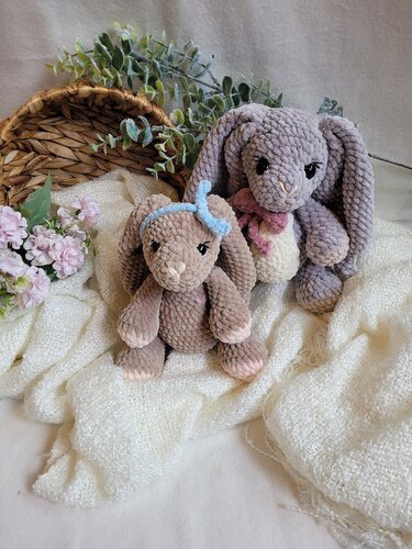 Zwei gehäkelte Amigurumi-Hasen aus Chenillegarn auf weißer Decke, einer mit blauer, einer mit rosa Schleife.