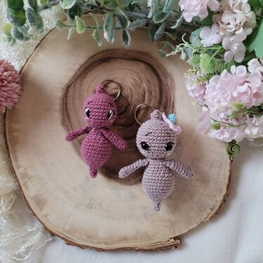 Zwei gehäkelte Amigurumi-Schlüsselanhänger in Rosé und Flieder auf einer Holzscheibe
