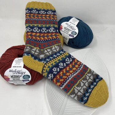 Gestrickte Socke mit buntem Fair-Isle-Muster und zwei Knäueln Wolle auf hellem, strukturiertem Untergrund.