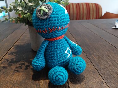 Türkis gehäkelte Amigurumi-Puppe mit roten Zierstichen, Knopf auf dem Kopf und rotem Halsband auf Holztisch