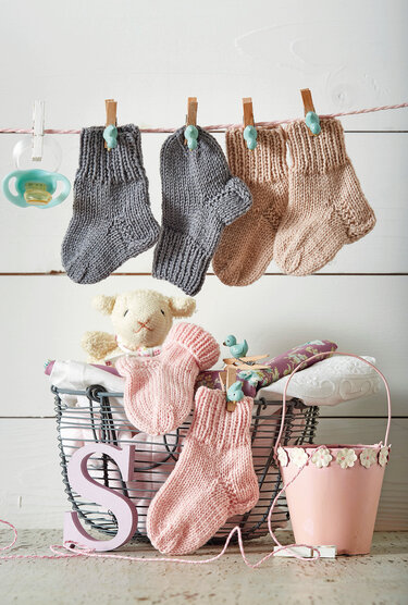 Gestrickte Babysocken in Grau und Rosa an einer Leine, darunter Drahtkorb mit Kuscheltier und weiteren Socken