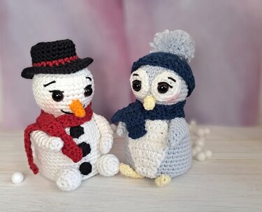 Gehäkelte Amigurumi: Schneemann mit Zylinder und rotem Schal neben Pinguin mit blauer Bommelmütze und Schal