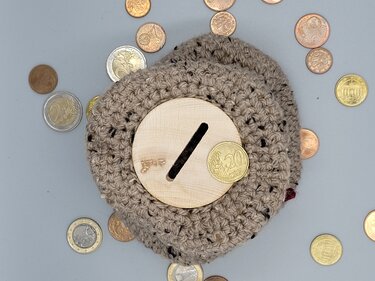 Beigefarbene gehäkelte Spardose mit runder Holzscheibe und Münzschlitz, umgeben von Euromünzen auf hellem Hintergrund.