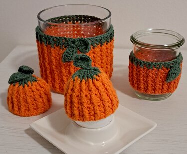 Zwei gehäkelte Kürbis‑Glaswärmer in verschiedenen Größen und ein gehäkelter Kürbis‑Eierwärmer, orange mit grünen Blattapplikationen.
