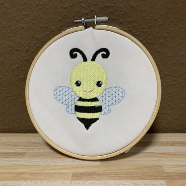 Gestickte gelb-schwarz gestreifte Biene mit hellblauen, geometrischen Flügeln in rundem Holzstickrahmen