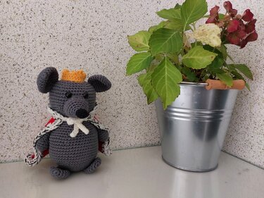 Gehäkelte graue Amigurumi-Maus mit gelber Krone und rotem Umhang neben Topfpflanze in Metallkübel