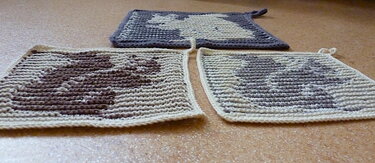 Drei gehäkelte quadratische Topflappen in Grau, Braun und Naturweiß mit stilisiertem Mausmotiv und Aufhängeschlaufe.