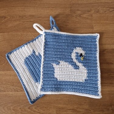 Zwei gehäkelte Topflappen in Blau-Weiß, einer mit Schwanmotiv, auf Holzuntergrund.