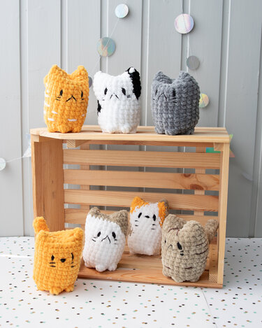 Sieben gehäkelte Katzen-Amigurumi in verschiedenen Farben auf einer Holzkiste.