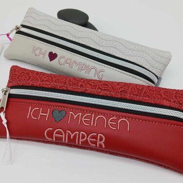 Zwei genähte Reißverschlusstaschen in Grau und Rot mit gestickten Schriftzügen 'ICH ♥ CAMPING' und 'ICH ♥ MEINEN CAMPER'.