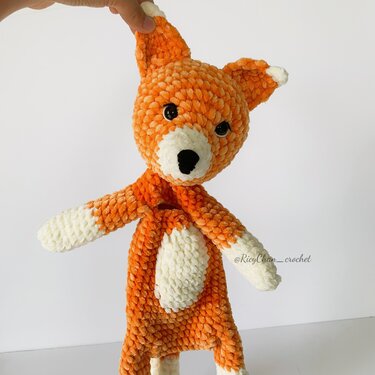 Hand-crocheted orange fox lovey toy with cream muzzle, belly, and paws held up against a white background