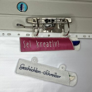 Zwei bestickte ITH-Stiftehalter zum Einhängen in Ordner, pink mit 'Sei kreativ!' und weiß mit 'Geschichten-Schreiber', jeweils mit Stift.