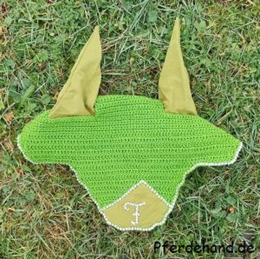 Grüne gehäkelte Fliegenmütze für Pferde mit Stoffohren und gesticktem „F“ auf Gras