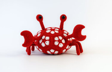 Rotes, gehäkeltes Amigurumi in Krabbenform mit weißen African-Flower-Motiven und schwarzen Augen auf weißem Hintergrund.