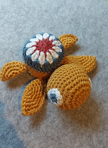Nahaufnahme einer gehäkelten Amigurumi-Schildkröte in Ocker mit blau-weiß-rotem Panzer und Blumenmuster auf grauem Hintergrund