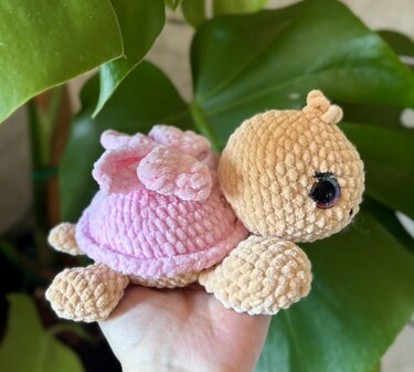 Hand hält kleine gehäkelte Amigurumi-Schildkröte aus Plüschgarn mit rosa Panzer und glänzenden Augen