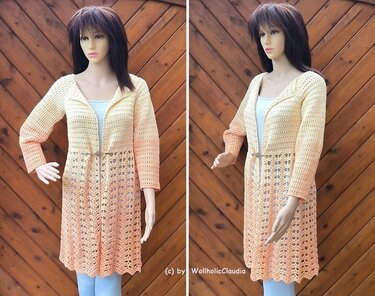 Mannequin trägt eine lange gehäkelte Longjacke in Creme‑Pfirsich-Ombré mit Lochmuster und Gürtel vor einer Holzverkleidung.