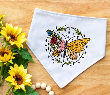 Weißes Stoffdreieck mit gesticktem Schmetterling, umgeben von buntem Blumenkranz