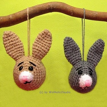 Zwei gehäkelte Hasenkopf-Anhänger, einer braun, einer grau, mit Schlaufe an einem Ast vor grünem Hintergrund