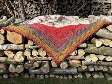 Gehäkeltes Dreieckstuch in Rot-, Orange- und Gelbtönen mit Strukturmuster auf einem Holzstapel.