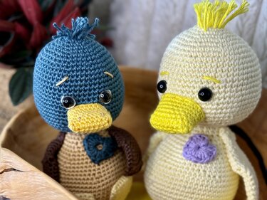 Zwei gehäkelte Entchen-Amigurumi, links blau/braun mit gelbem Schnabel, rechts cremefarben mit gelbem Schnabel und lila Herz