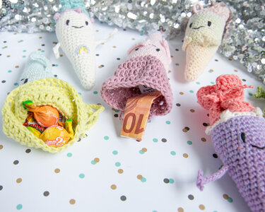 Mehrere schnell gehäkelte Amigurumi-Täschchen in Pastellfarben, einige geöffnet und mit Süßigkeiten oder Geldscheinen gefüllt, auf festlichem Konfetti-Hintergrund.