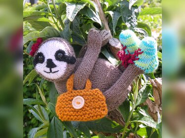 Gestricktes Faultier-Amigurumi in Braun mit weißem Gesicht, orangefarbener Tasche und türkis-grünen Füßchen, hängend an einem Ast