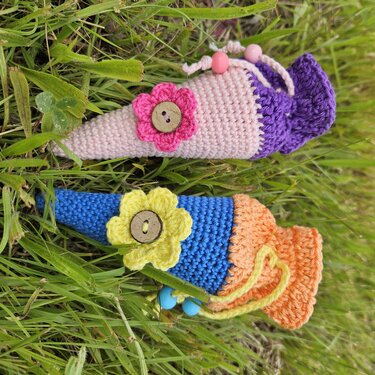 Zwei gehäkelte Mini-Schultüten in Rosa/Violett und Blau/Orange mit Blumenapplikationen, Holzknöpfen und Perlenband auf Gras