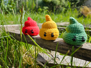 Drei gehäkelte Amigurumi-Emojis (rot, gelb, grün) mit mürrischem Gesicht auf einem Holzbrett im Gras.