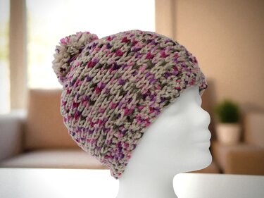 Gehäkelte Bommelmütze in Beige mit pinken und lila Sprenkeln auf weißer Schaufensterpuppe