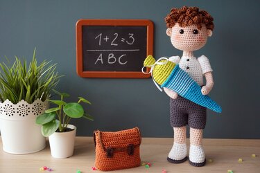 Gehäkelter Amigurumi-Junge in weißem Hemd und dunkler Hose, der eine blau-gelbe Schultüte hält; im Hintergrund eine Tafel mit '1+2=3' und 'ABC' sowie ein gehäkelter Ranzen.