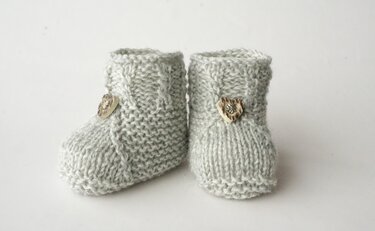 Ein Paar graue, gestrickte Babyschuhe mit herzförmigen Knöpfen auf weißem Hintergrund.
