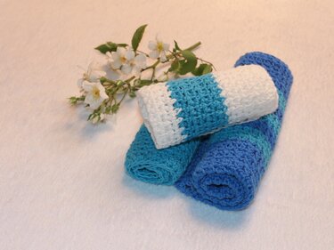 Drei gerollte gehäkelte Tücher in Blaunuancen und Weiß liegen auf hellem Untergrund neben einem kleinen Blumenzweig.