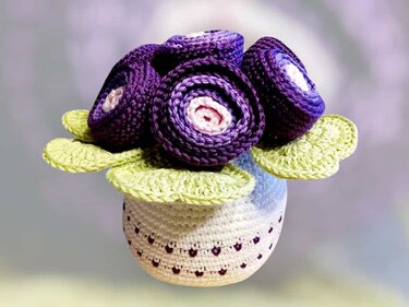 Weiß gehäkelter Topf mit grünen Blättern und drei lila-rosa gehäkelten Rosen