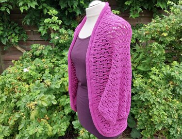 Magenta gestrickter Seelenwärmer mit Ajourmuster auf einer Schaufensterpuppe vor Grünpflanzen