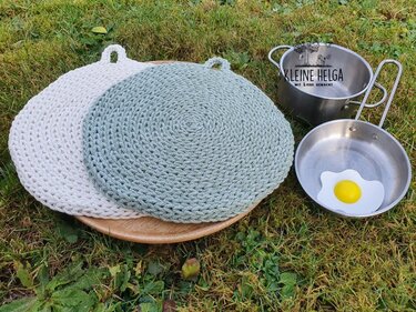 Zwei runde gehäkelte Topflappen in Beige und Mint auf einer Holzplatte im Gras, daneben Spielzeug-Küchenutensilien.