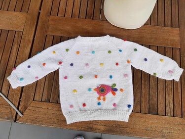 Weißer gestrickter Baby-Pullover mit bunten Noppen und bunter Vogel-Applikation, liegend auf Holztisch