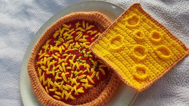 Gehäkelte Spiel-Pizza mit gelben Käsefäden und brauner Kruste neben quadratischer gelber Käsescheibe